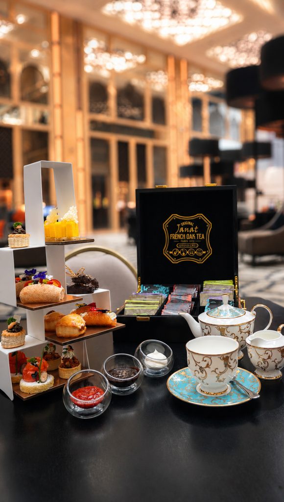 A tiered stand of finger sandwiches, scones and pastries beside a teapot, a china cup and a box of French black tea on a dark table