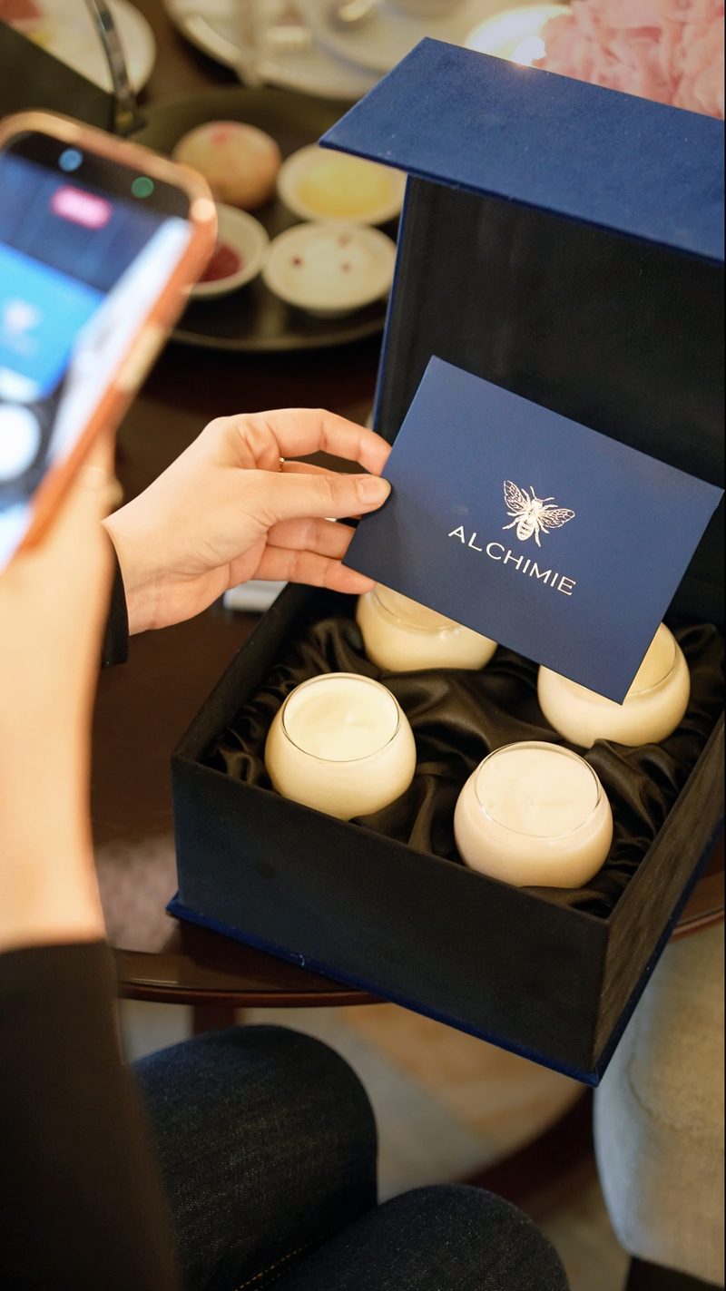 A hand holds an Alchimie branded card over an open box containing candles, with a phone photographing it