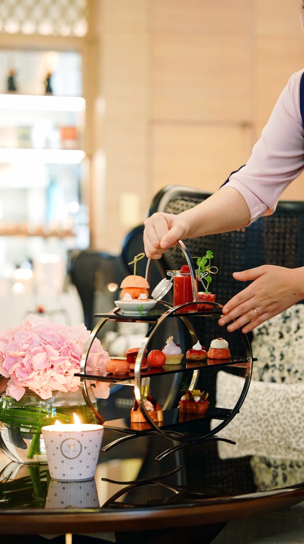 A hand places a dessert on a tiered stand with pastries, beside a pink flower arrangement and candle
