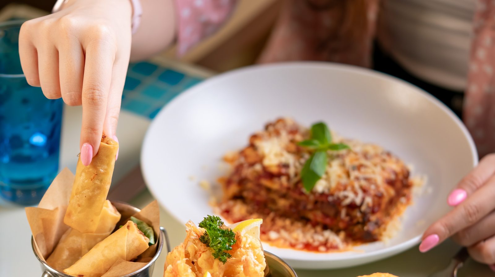 A hand reaching for a crisp roll from a metal pot of bar bites, beside a plate of baked pasta topped with a basil leaf