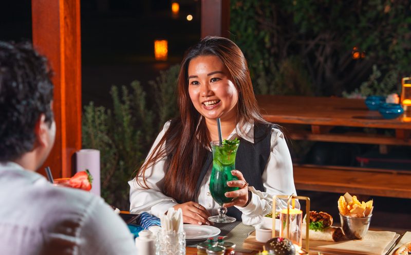A smiling woman holding a green cocktail at an outdoor evening table set with sharing plates and fries