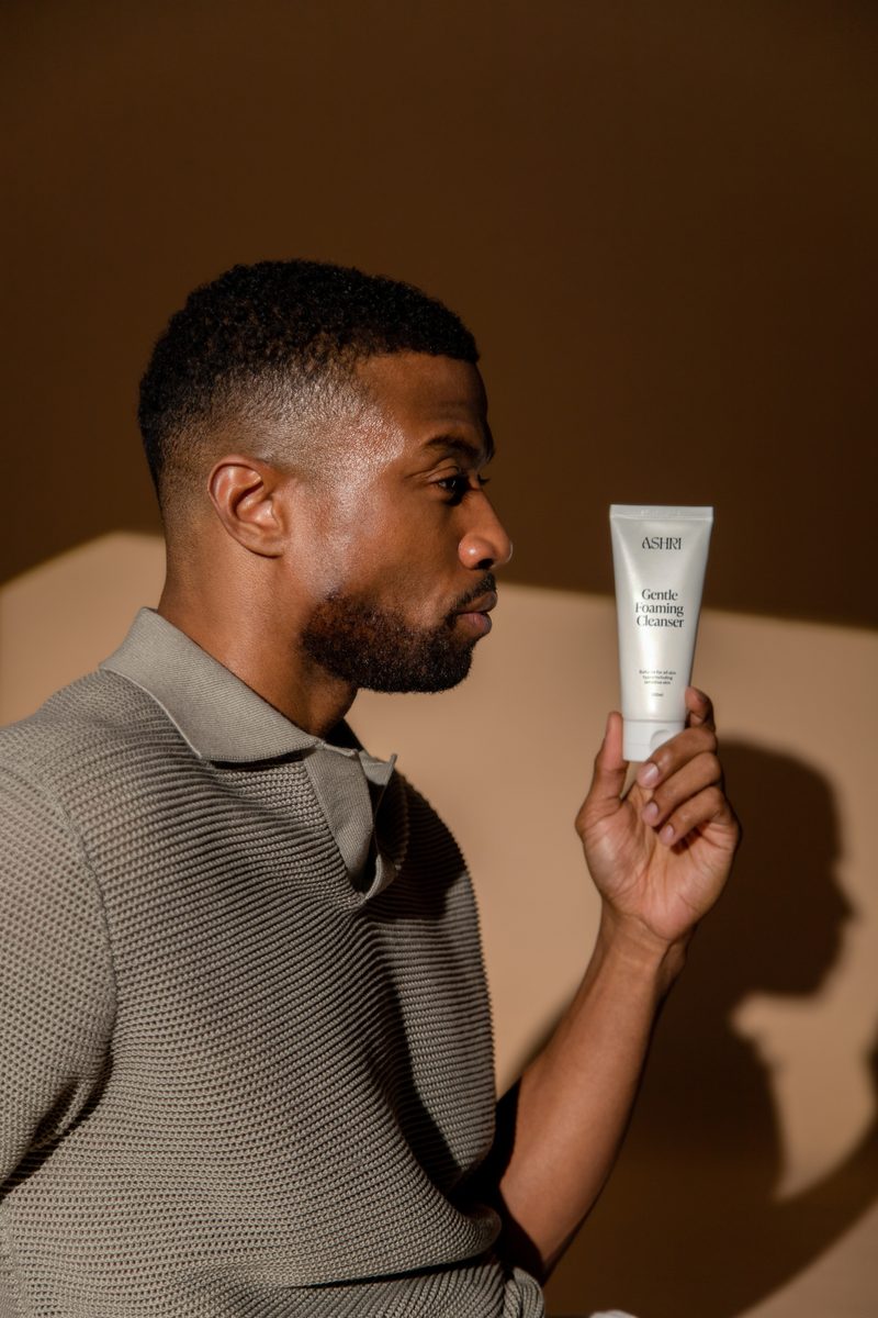 A man in a grey knit polo holds up a tube of Ashri Skin product against a warm brown background