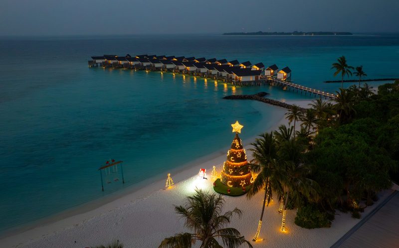 A lit Christmas tree on the beach beside overwater villas at dusk at an Atmosphere resort