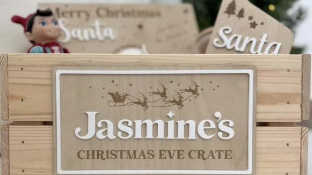 A personalised wooden Christmas Eve crate with a sleigh-and-reindeer nameplate, a door hanger and an elf toy in front of a Christmas tree