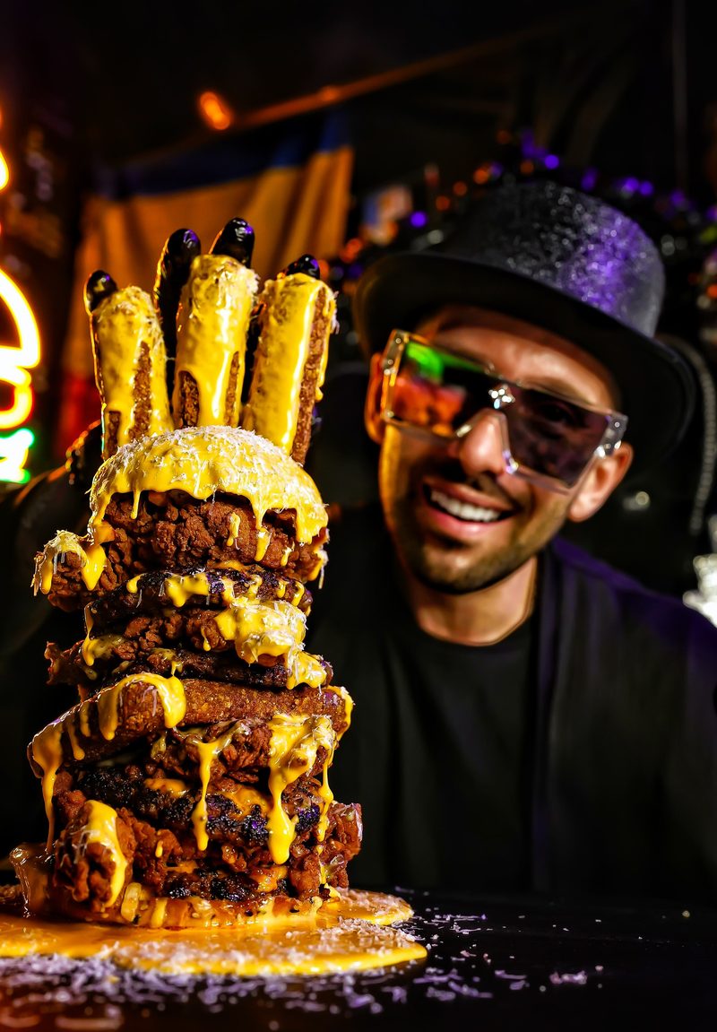 Owner Alin Ciobanu in a top hat and tinted glasses beside a tall stack of burger patties and fries