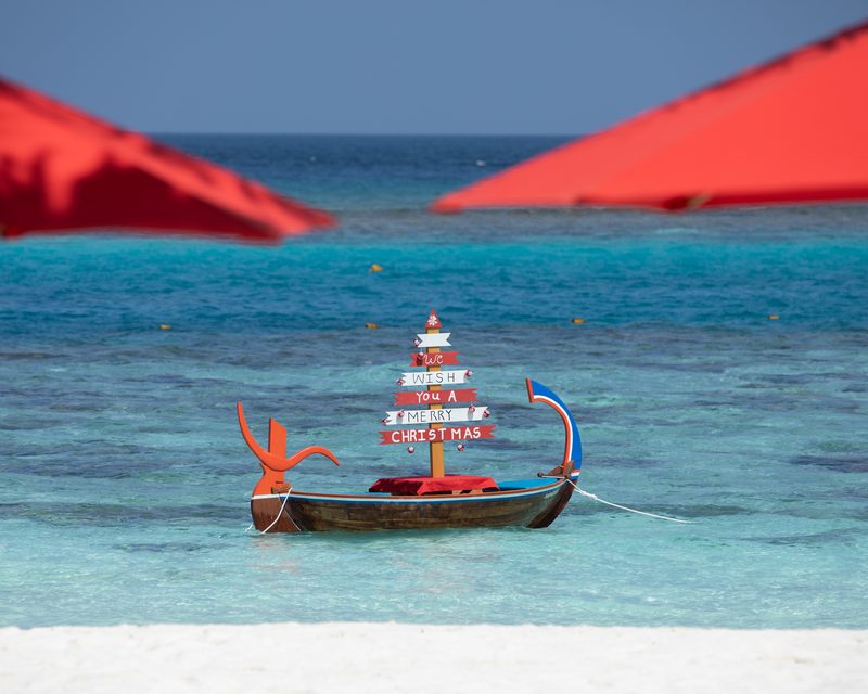 A traditional wooden Maldivian dhoni in shallow water carrying a signpost that reads We wish you a Merry Christmas, between two red umbrellas