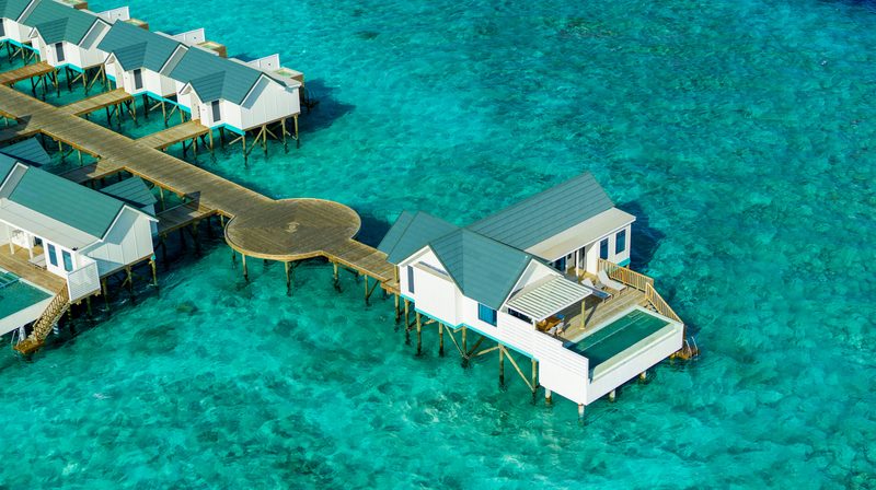 Aerial view of white overwater villas with grey roofs, one with a private pool, along a wooden walkway over turquoise water