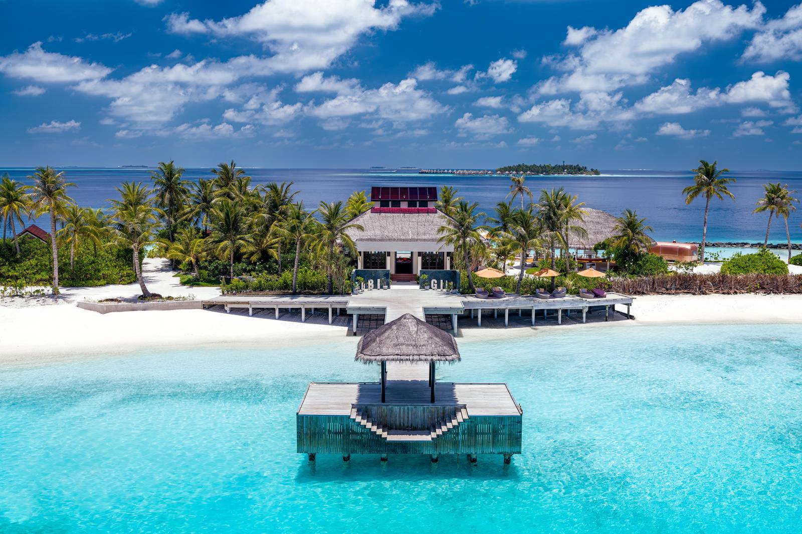 Aerial view of a jetty and thatched pavilion over turquoise water, with a beachfront building among palm trees behind