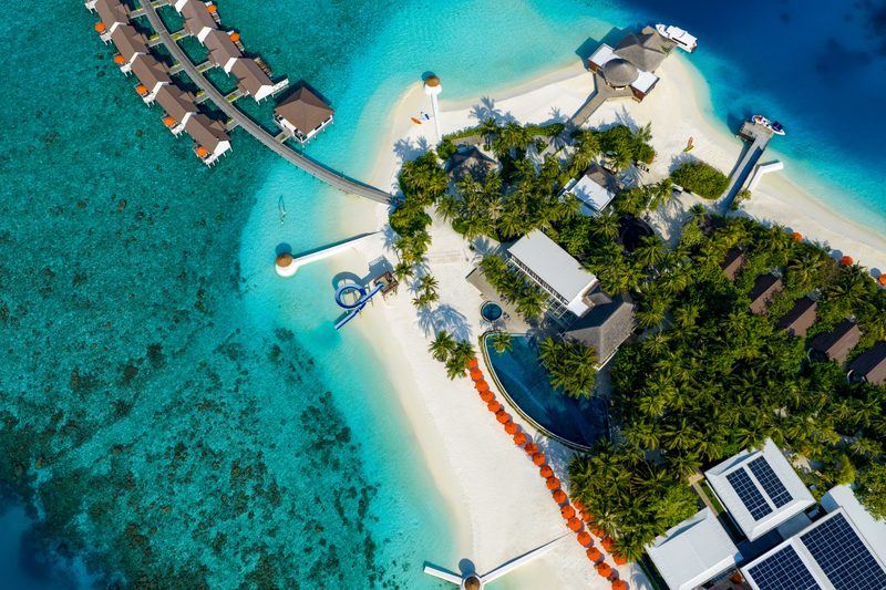 Aerial view of a palm-covered island with white beaches, overwater villas along a long jetty and a blue water slide