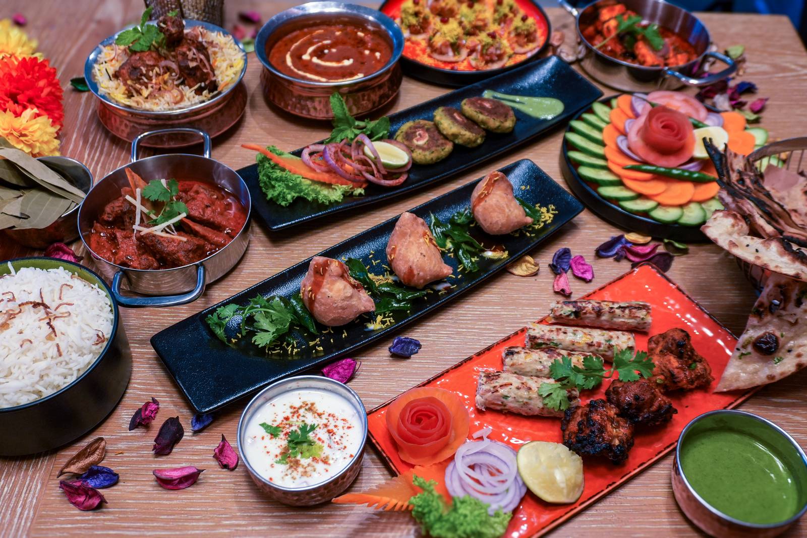 Overhead view of Indian dishes including kebabs, curries, rice and salads on a wooden table decorated with marigolds