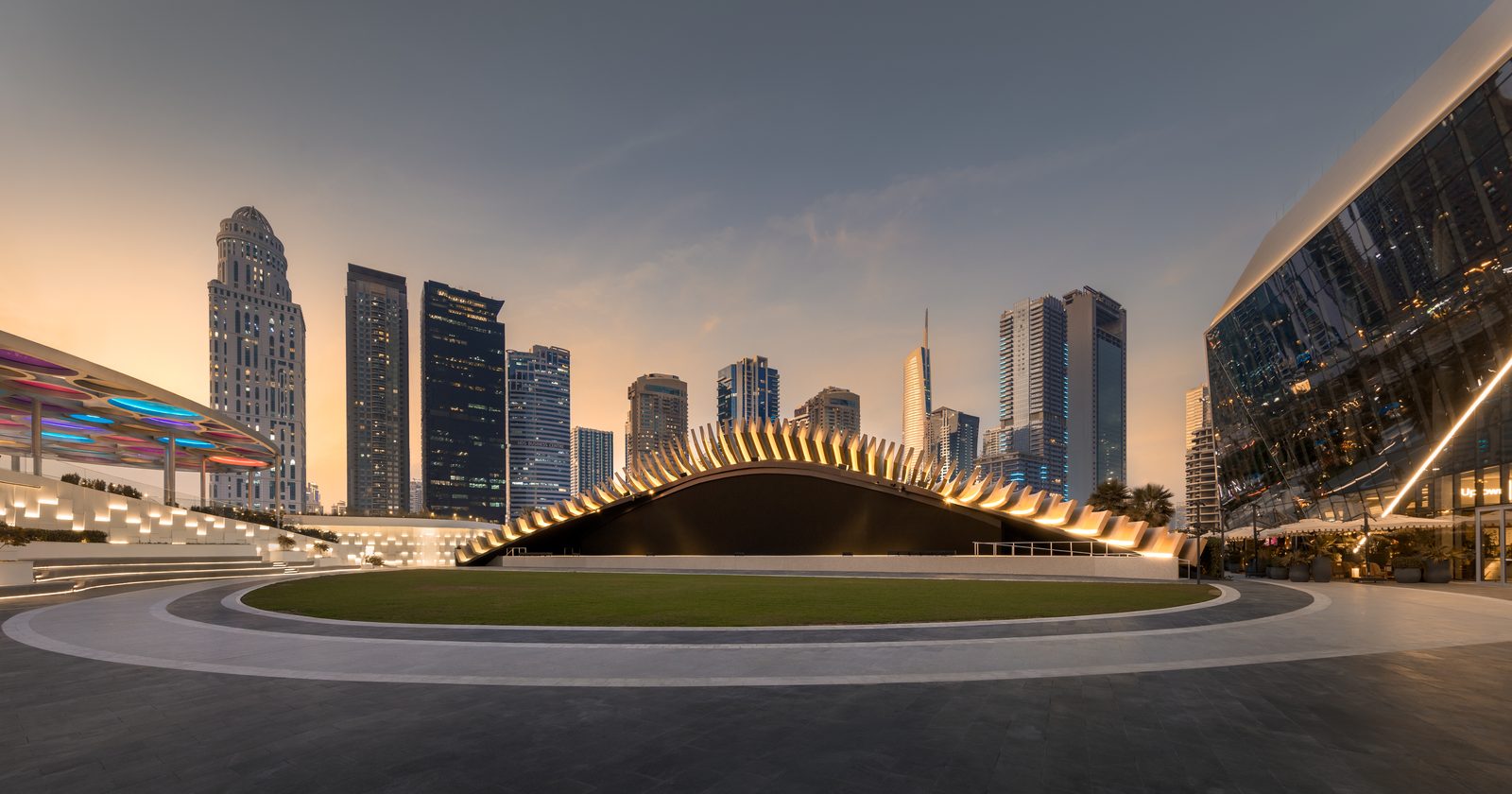 The Plaza at Uptown Dubai at sunset, with the lawn and curved stage roof in front of the Dubai skyline