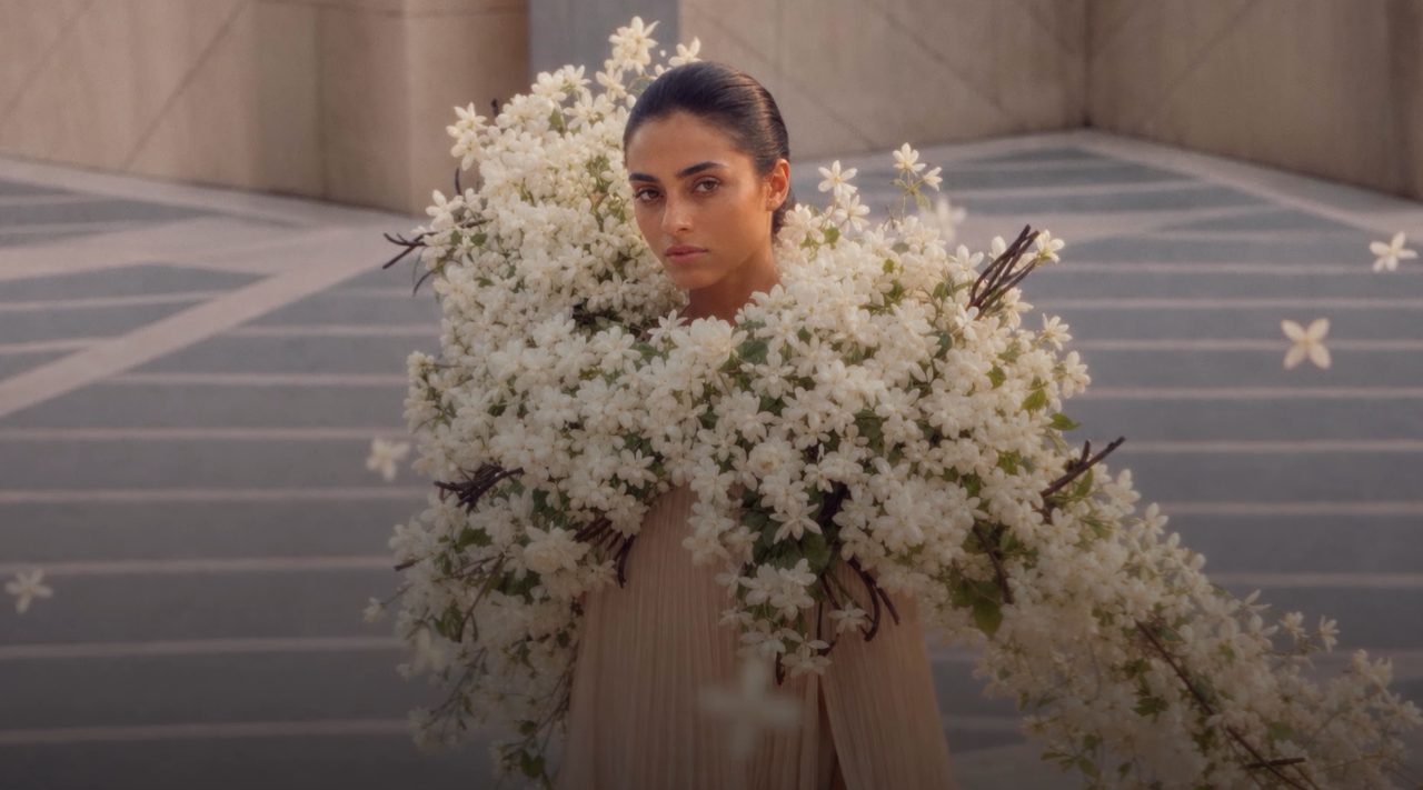A model with her hair tied back looks at the camera on a paved terrace, wearing a pleated cream cape with a large arrangement of white flowers across her shoulders