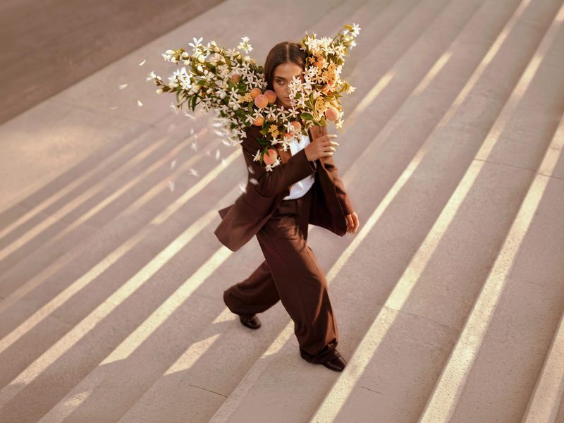 A model in a brown suit walks down wide stone steps carrying a large bouquet of white flowers on her shoulder, with petals in the air around her