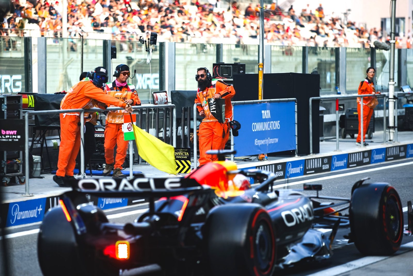Track marshals in orange overalls with a flag beside a Formula One car in the pit lane
