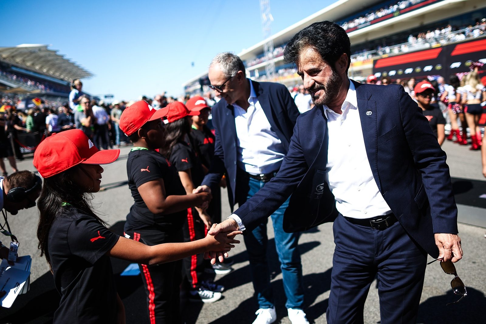 FIA President Mohammed Ben Sulayem shaking hands with a young person in a red cap among a crowd on a racetrack grid