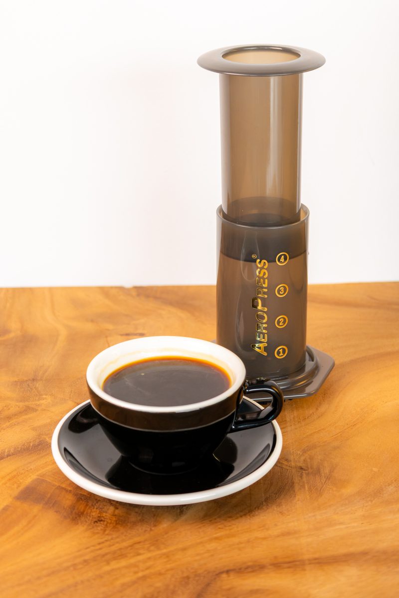 A black cup of coffee on a saucer beside an AeroPress on a wooden table