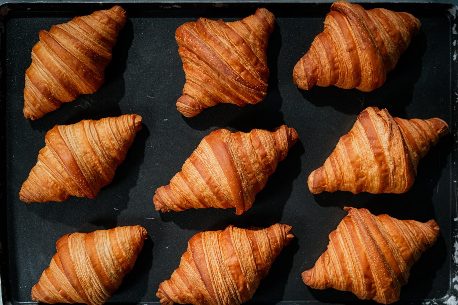 Nine freshly baked croissants on a dark baking tray at From Scratch by The Coffee Lab