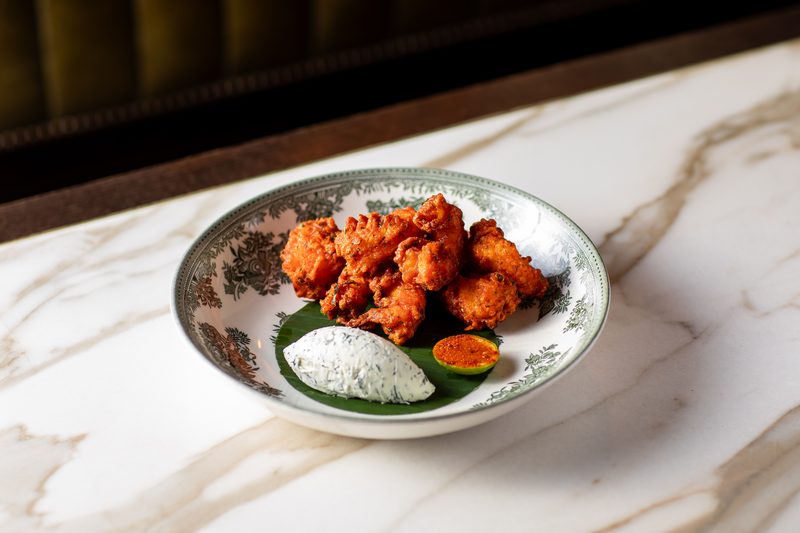 Fried battered shrimp in a patterned bowl on a leaf with a small pot of white chutney and a dipping sauce, on a marble table
