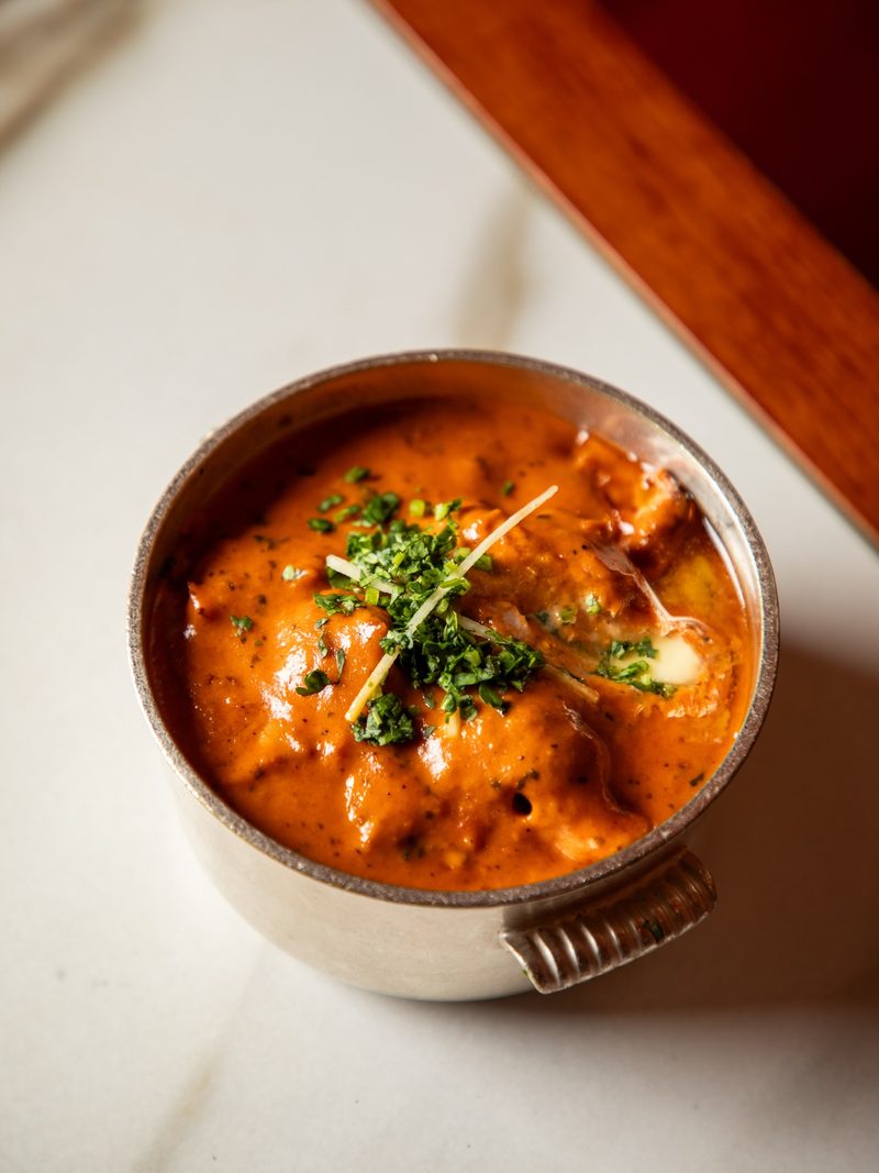 Metal pot of orange chicken butter masala garnished with herbs and ginger strips, on a marble surface