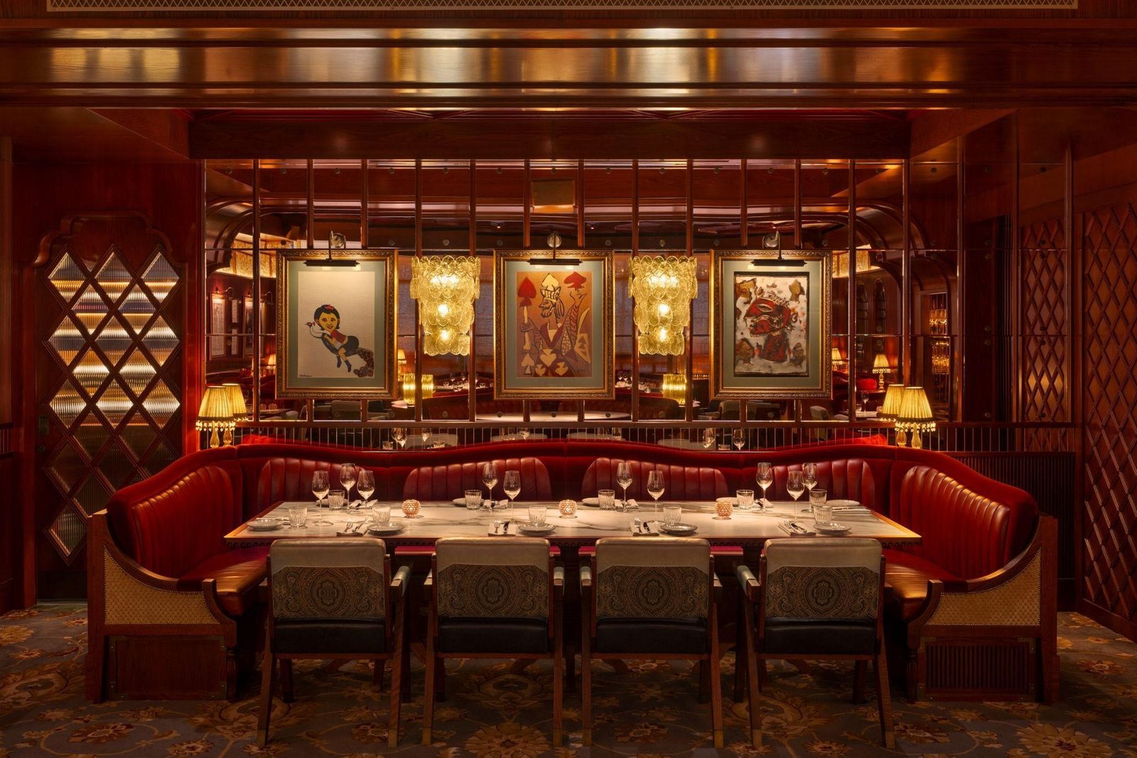 A long dining table laid with glasses and plates in front of red leather banquette seating, with three framed illustrations on the wall behind, at Gymkhana Dubai