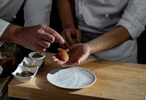 A chef's hands shaping a piece of sushi at a wooden counter, with small bowls and a white plate beside it