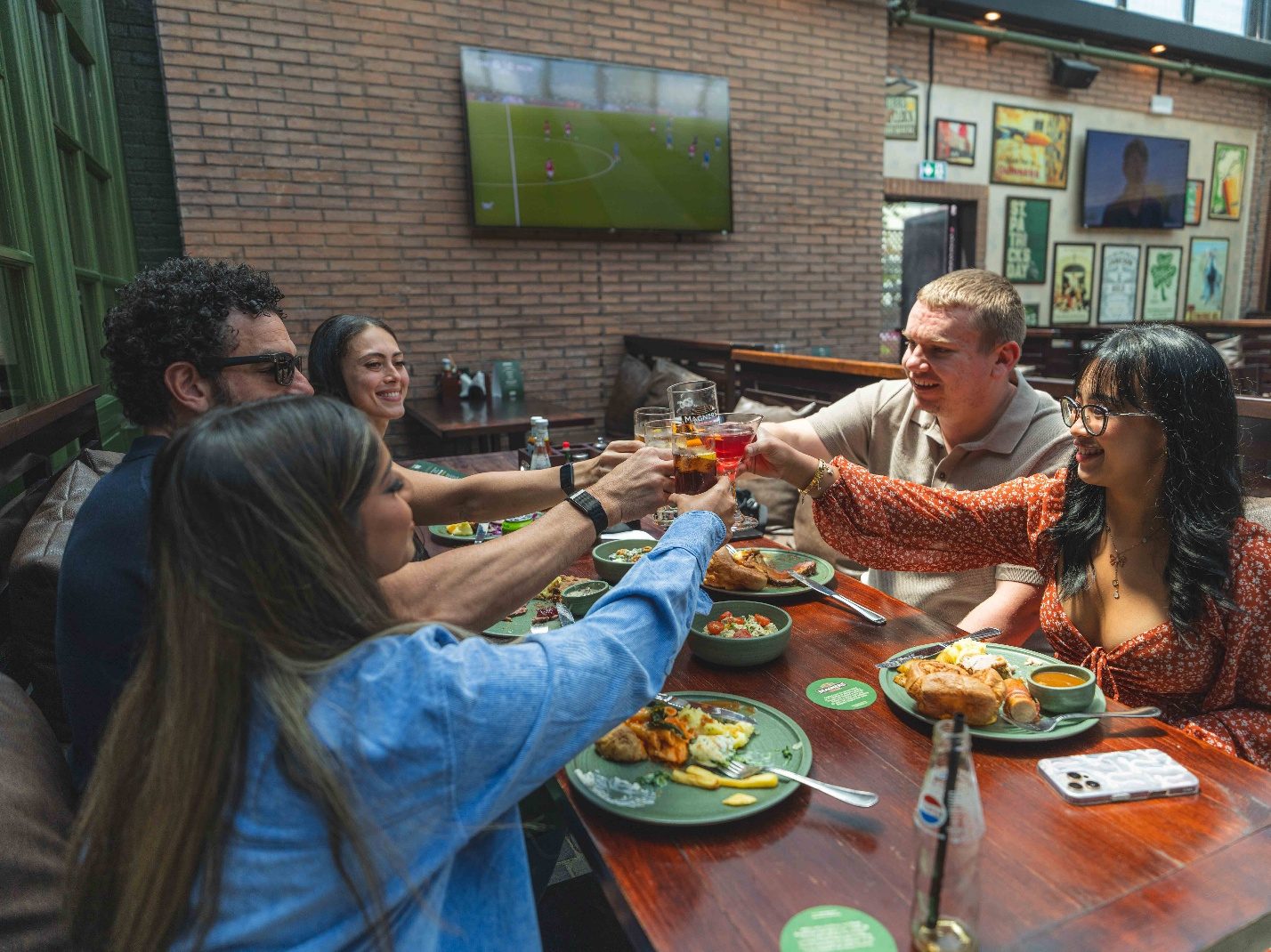 A group of friends raising glasses across a pub table laid with food, with a match on the screen behind them