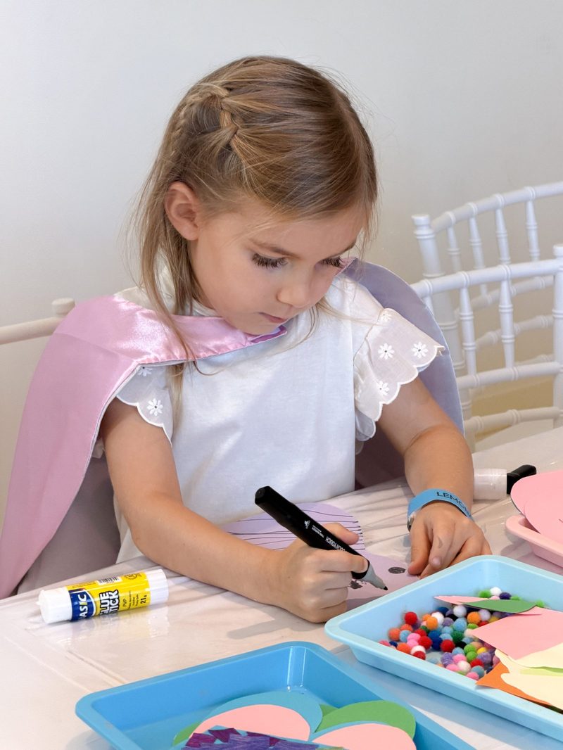 A girl in a pink cape drawing at the craft table at Lemonade Play