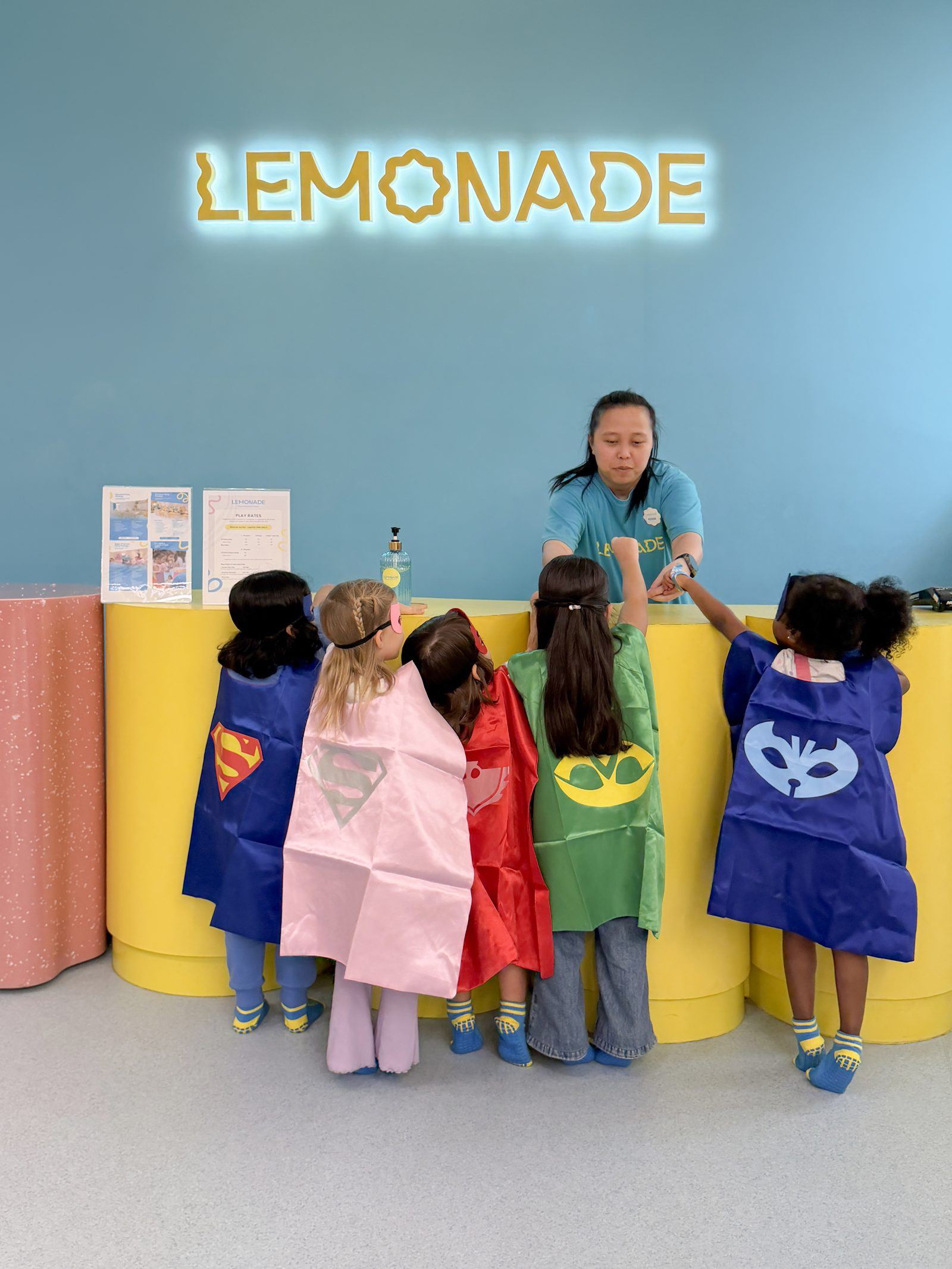 Children in superhero capes at the Lemonade counter in a Lemonade Play branch, with a member of staff