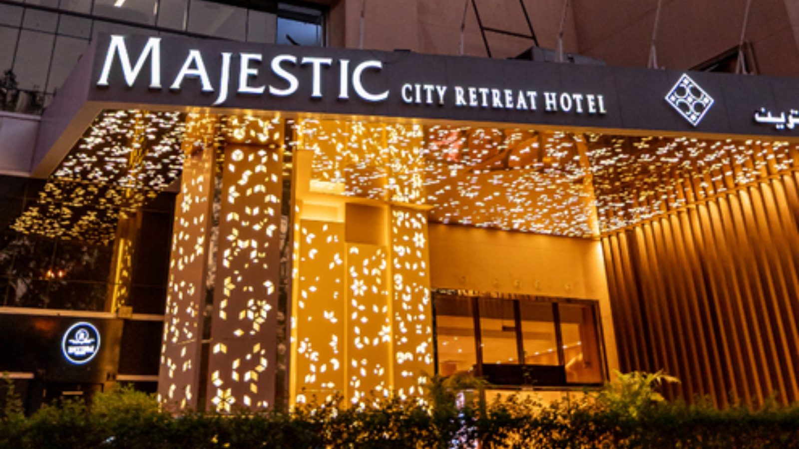 The lit entrance canopy of Majestic City Retreat Hotel at dusk, with the hotel name above the door and flags beside it