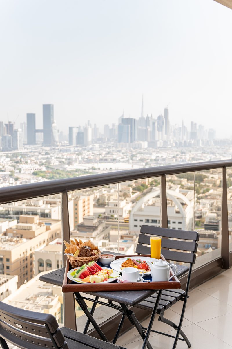 A breakfast spread on a small balcony table with the Dubai skyline in the haze behind