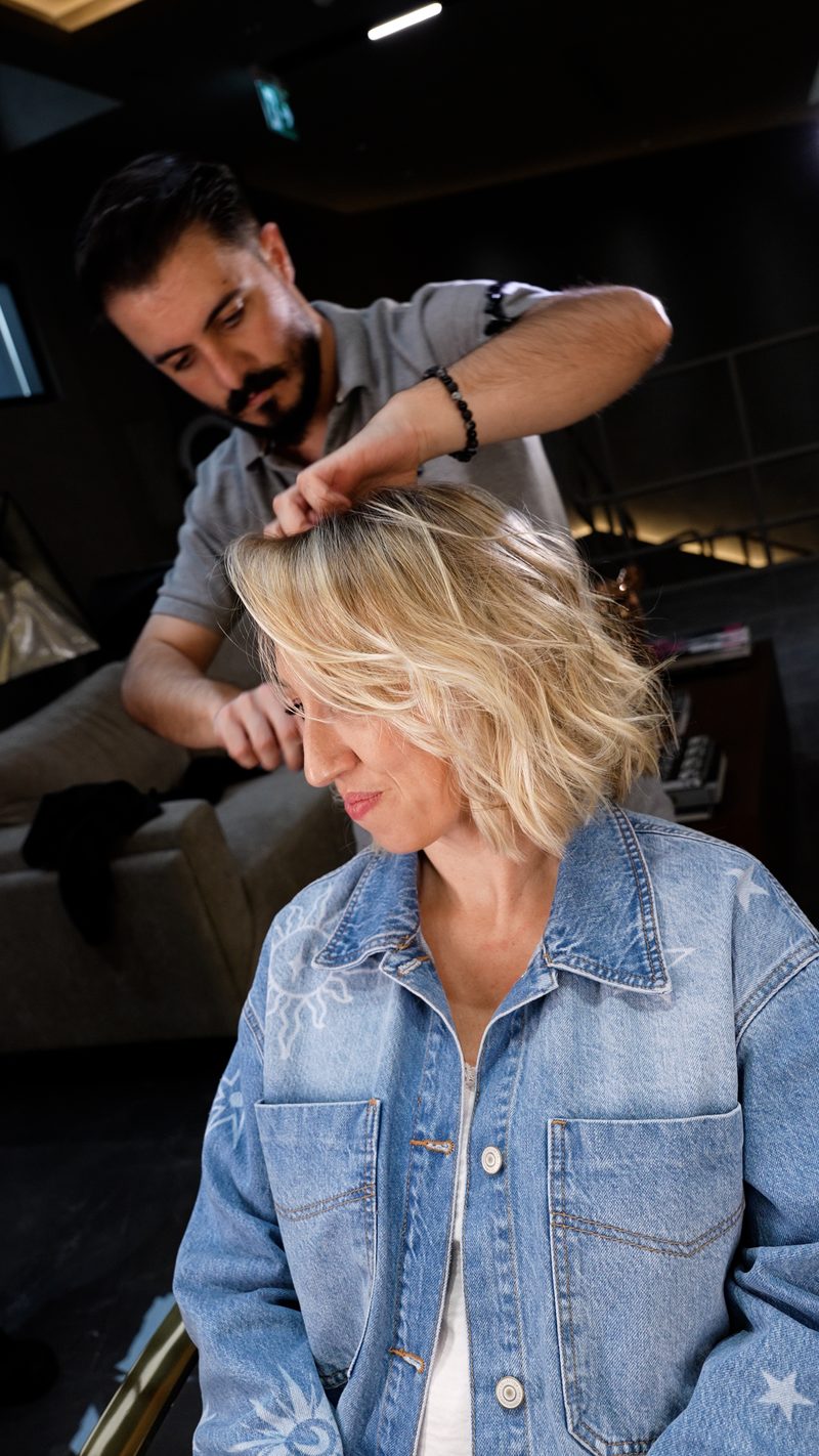 A stylist working on a client’s short blonde hair in a salon, in a denim jacket