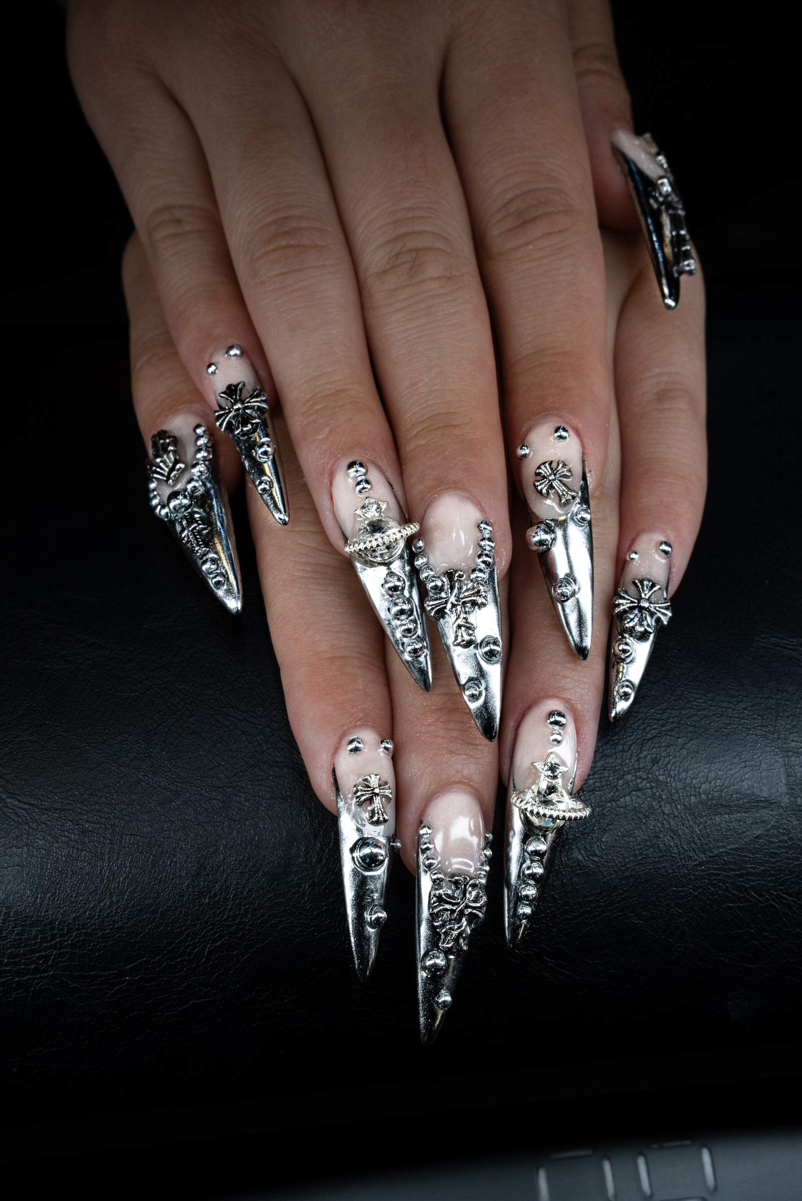Hands with long almond nails in chrome silver and black with raised silver cross decorations on a black leather surface
