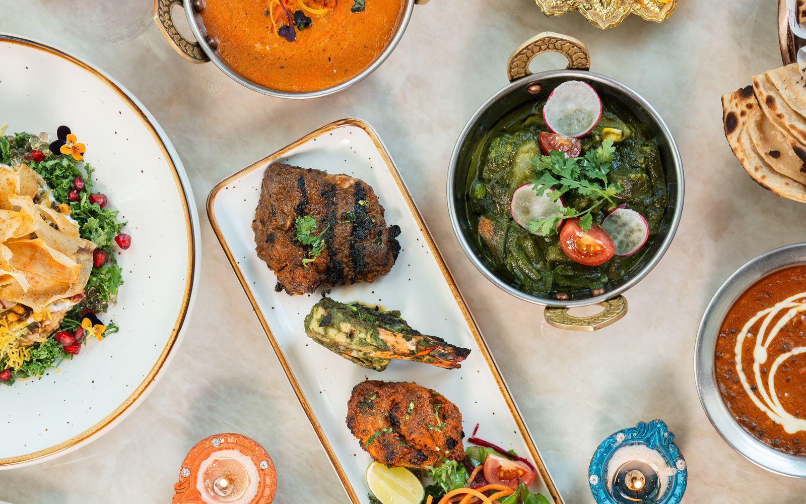 Overhead view of Indian dishes at Mausam, including kebabs on a white platter, a green curry, salad and lit diyas