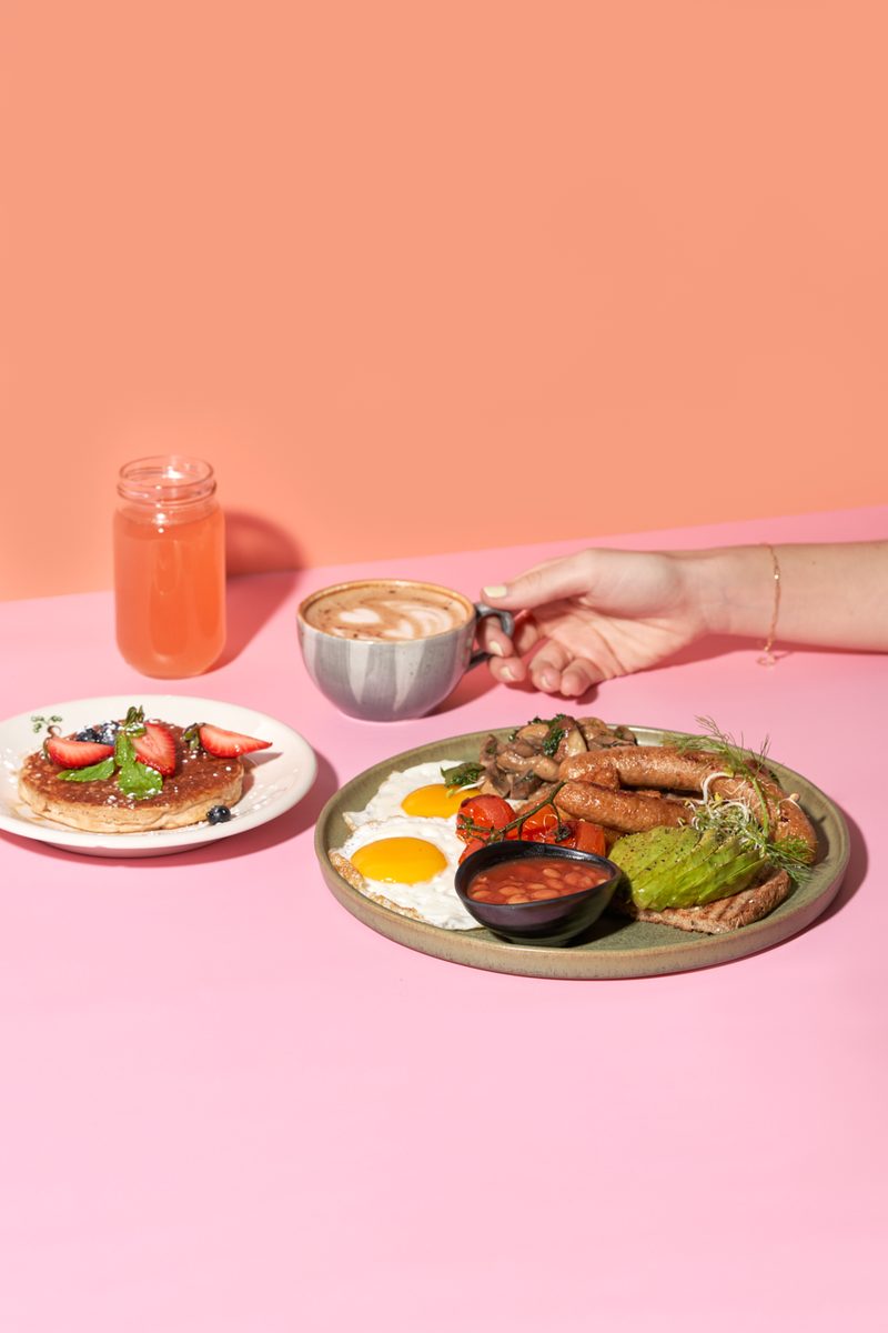 A breakfast set with a pancake, juice and a latte on a pink table at Organic Foods & Café