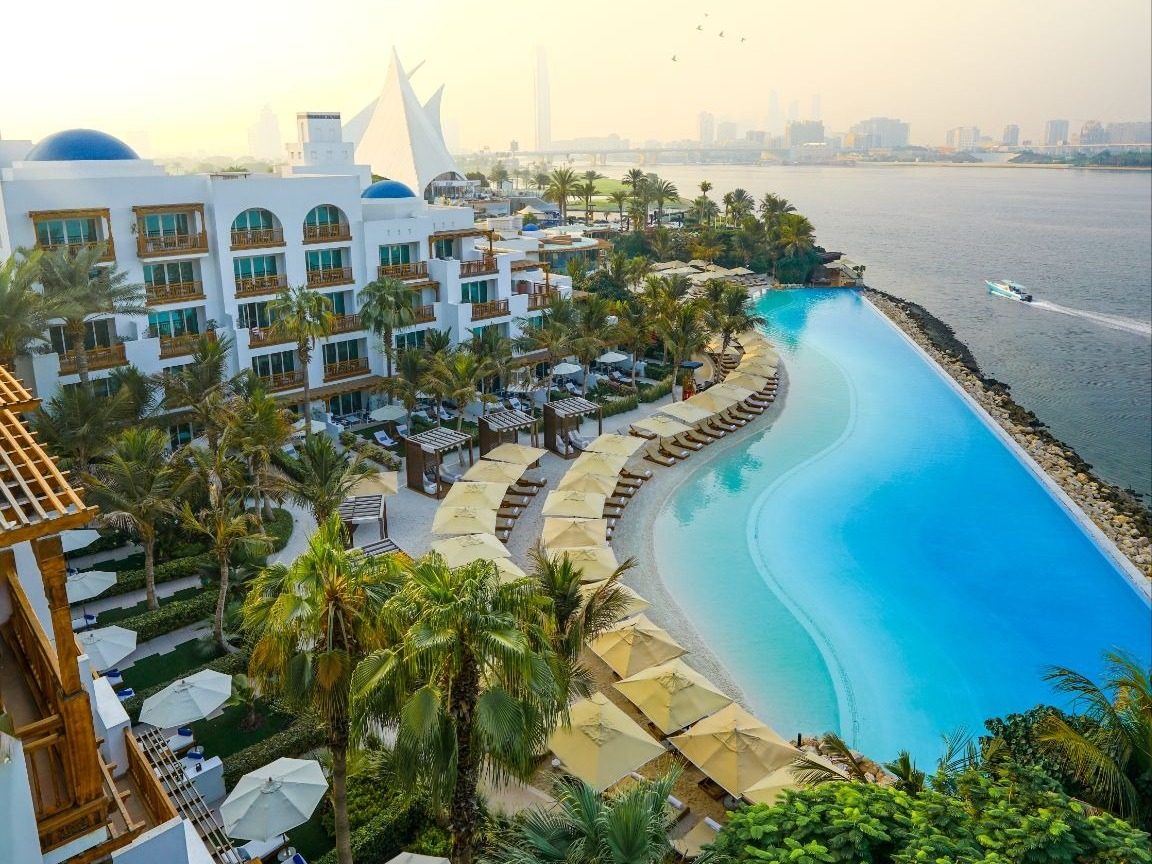 Aerial view of Park Hyatt Dubai, with white buildings, blue domes, a pool and palm gardens beside Dubai Creek