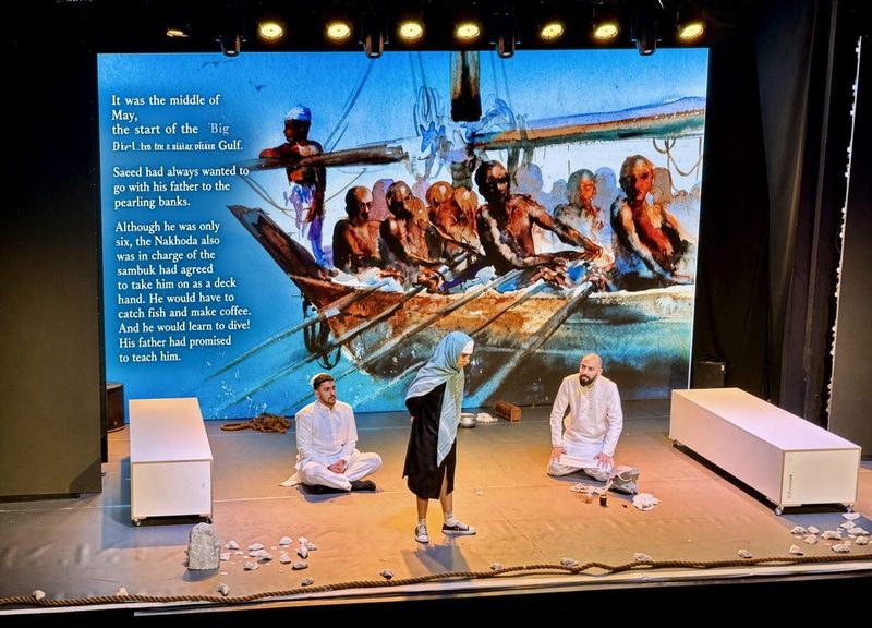 Three actors kneeling on stage in front of an illustrated boat backdrop in The Pearl Diver