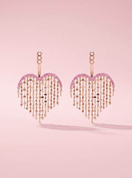 A pair of heart-shaped rose gold jacket earrings with pink sapphire strands on a pink background