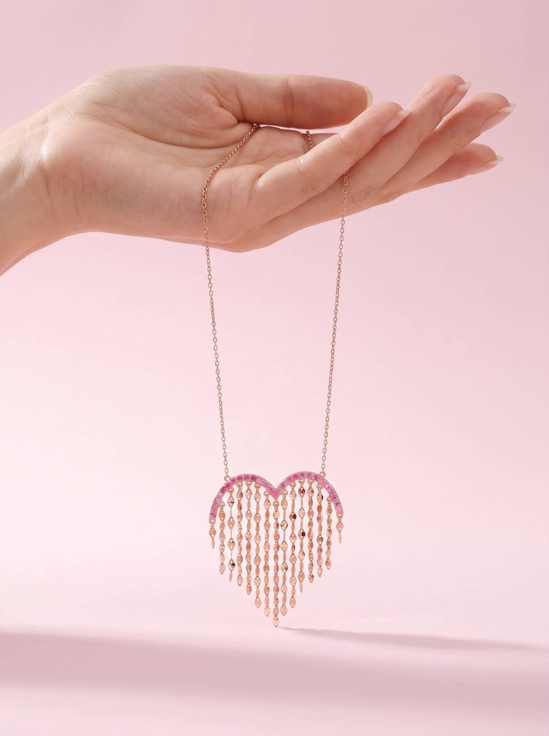 A hand holding up the Heartstring necklace by its chain against a pink backdrop