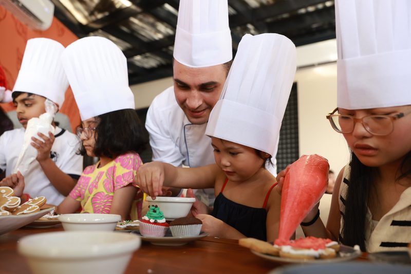 Children in chef hats decorating food with a chef at a long table