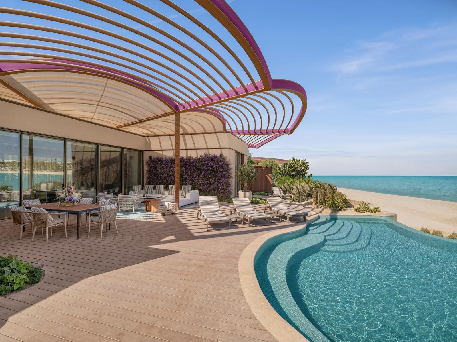 A resort terrace with a curved slatted canopy, lounge chairs and a curved swimming pool beside a sandy beach and the sea