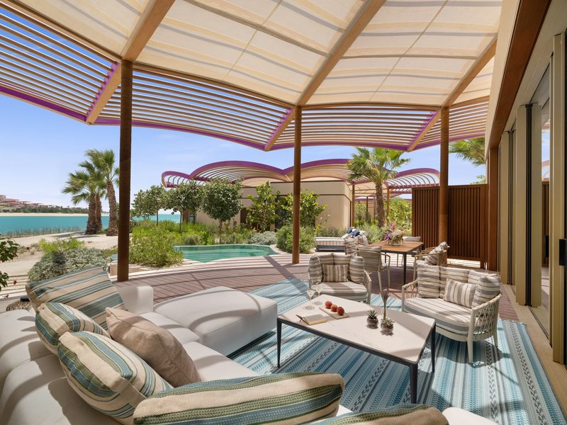 A shaded villa terrace with striped sofas, a coffee table and a small plunge pool, with palm trees beyond