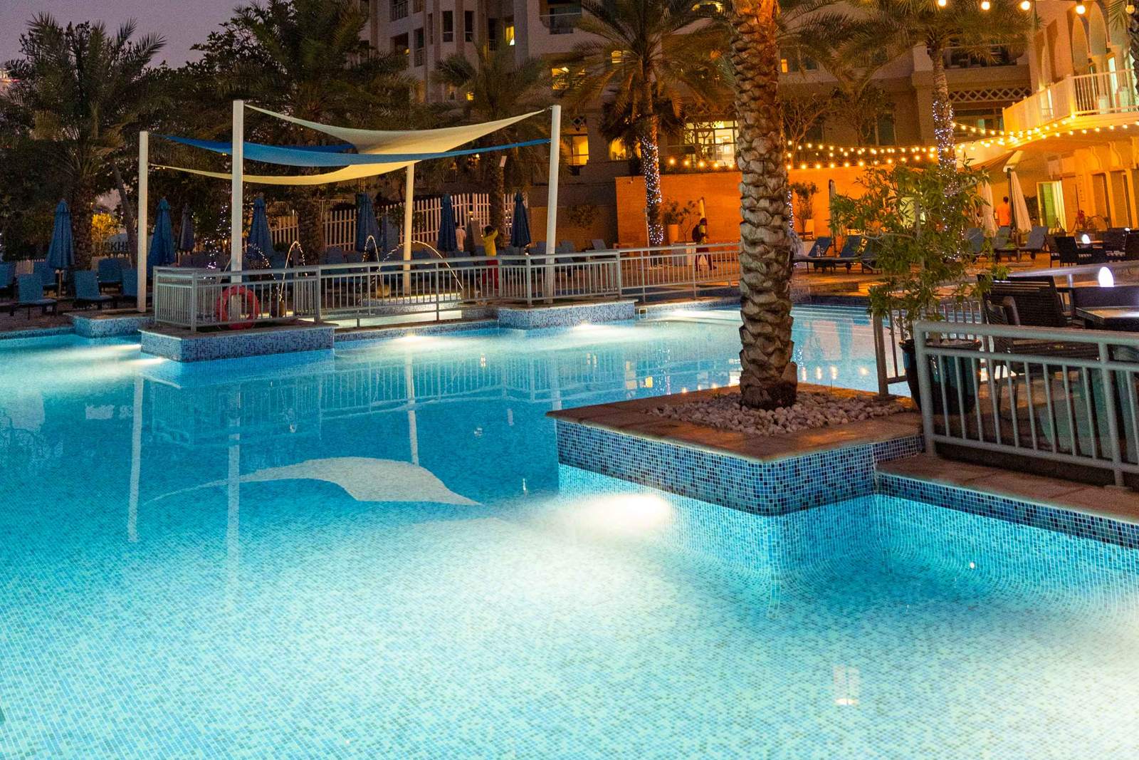 The pool at Riva Beach Club on Palm Jumeirah at dusk, with a shade sail, a palm tree and lit buildings behind