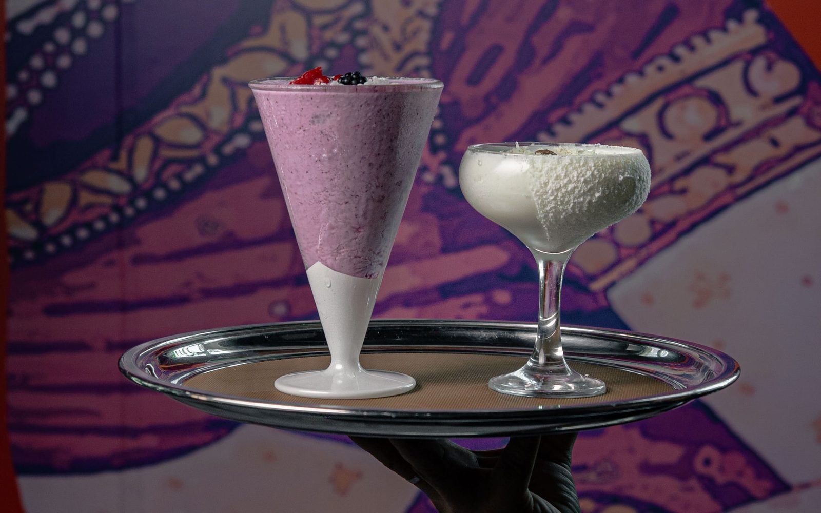 A pink berry lassi in a conical glass and a white khoya and coconut lassi in a coupe glass on a silver tray against a purple patterned mural