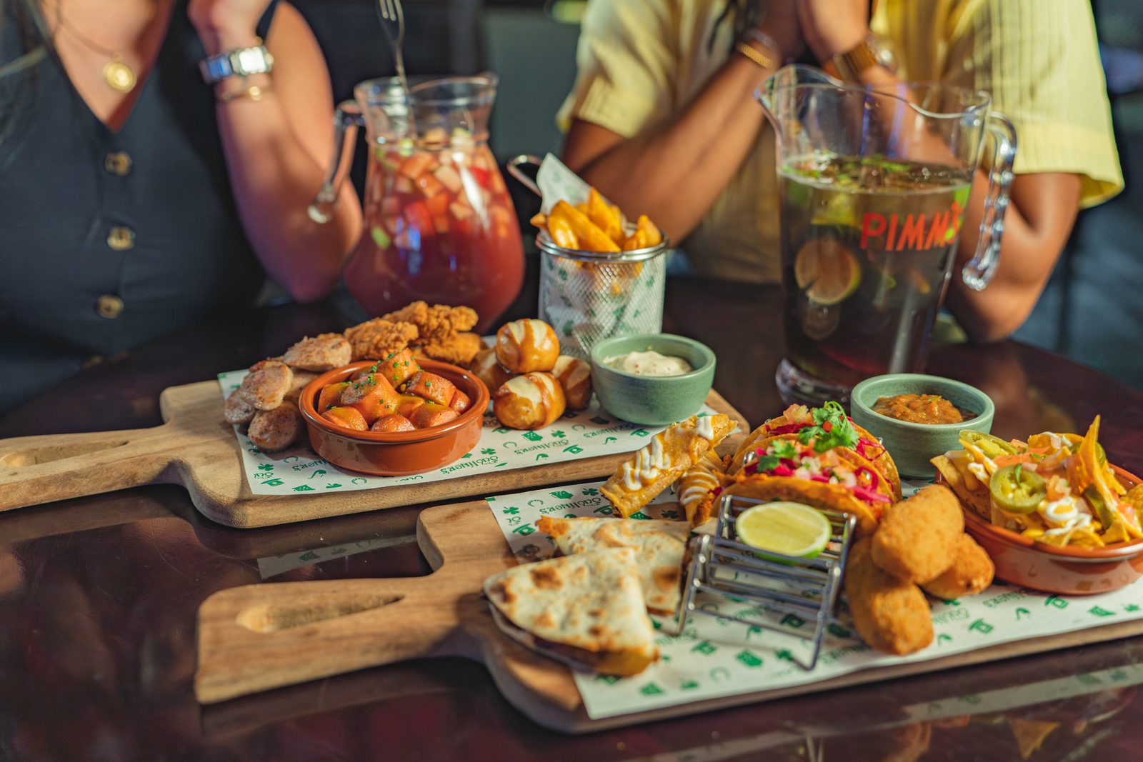 Sharing boards of fried chicken, taquitos, croquettes and fries on a pub table, with a jug of sangria and a jug of Pimm’s and two diners behind