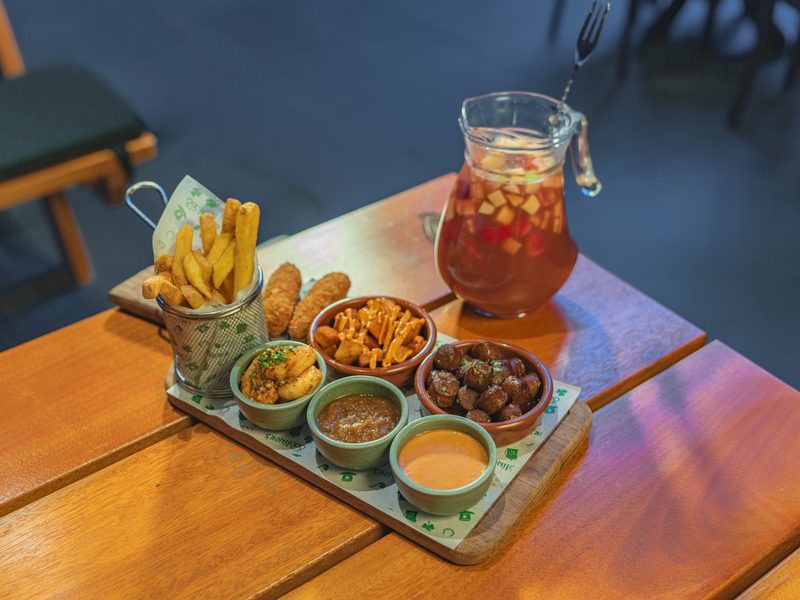 A wooden tray of fries, croquettes, sausage bites and dips with a jug of fruit sangria on a wooden pub table