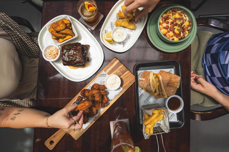 Overhead view of a pub table laid with ribs, potato wedges, fish bites, loaded potato, a toasted sandwich, fries and chicken wings on Le Méridien paper