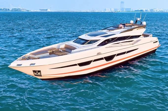 A cream-coloured motor yacht with an orange stripe along the hull on bright blue water, with a city skyline on the horizon