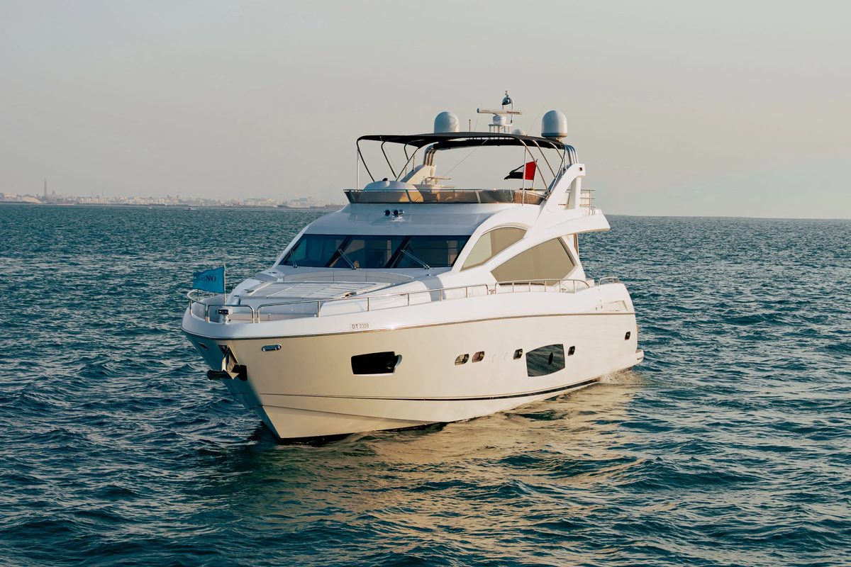 A white motor yacht seen from the front on dark blue open water, with a hazy coastline behind