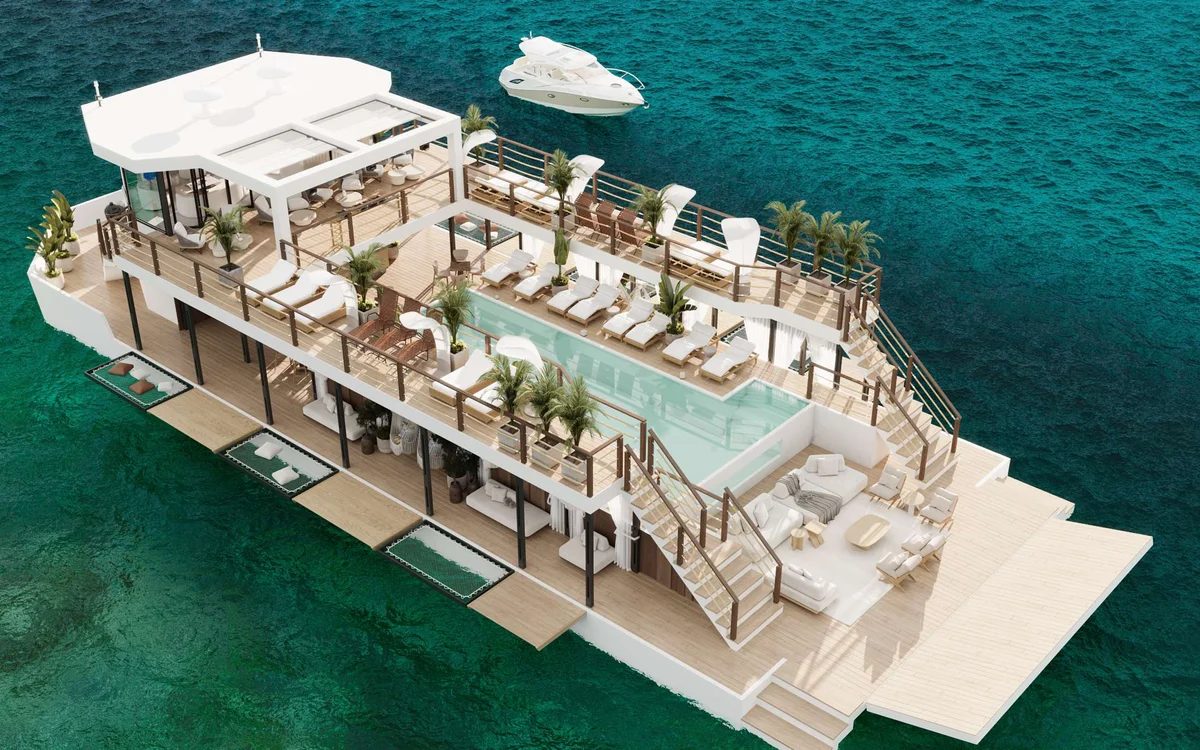 An aerial view of a large white multi-deck vessel with a swimming pool, sun loungers and palm trees on the upper deck, on turquoise water