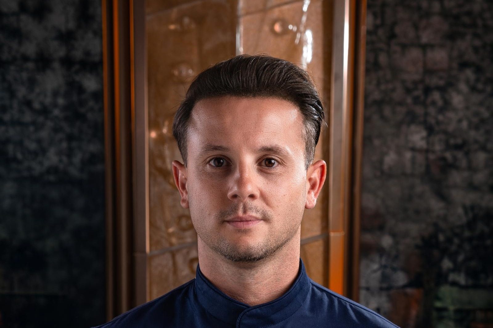A man in a navy chef jacket with the Sexy Fish Dubai name embroidered on the chest, in front of a dark wood and amber lit interior